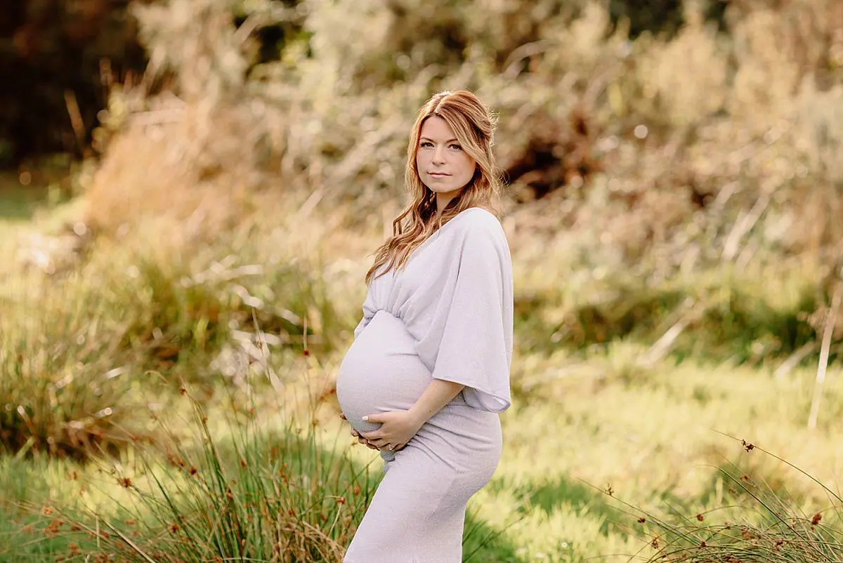 Expecting mother in a flowy white gown posing in a serene meadow during a maternity photoshoot by Ania Photo