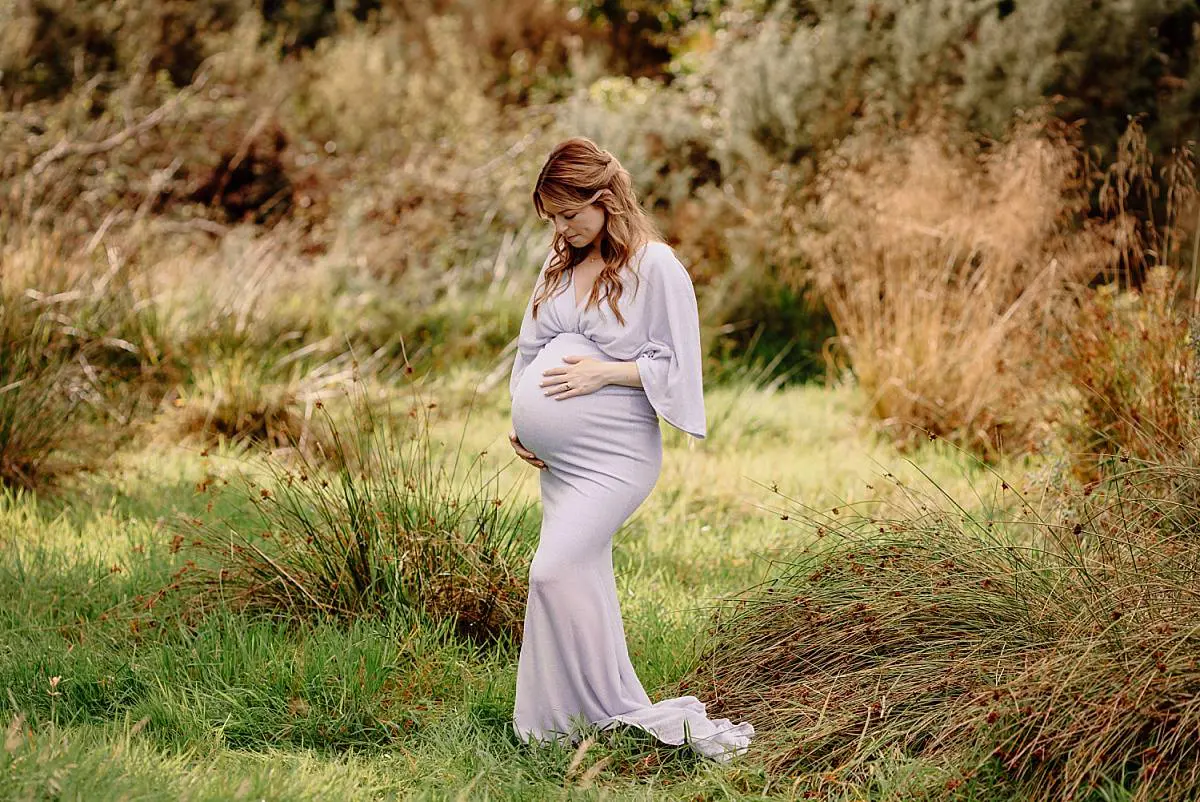 Expecting mother in a flowy white gown posing in a serene meadow during a maternity photoshoot by Ania Photo in Denmead