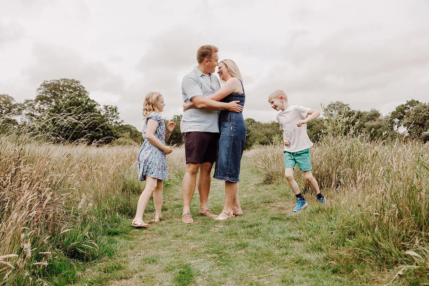 Parents hugging in field while children play during relaxed outdoor family session in Hampshire