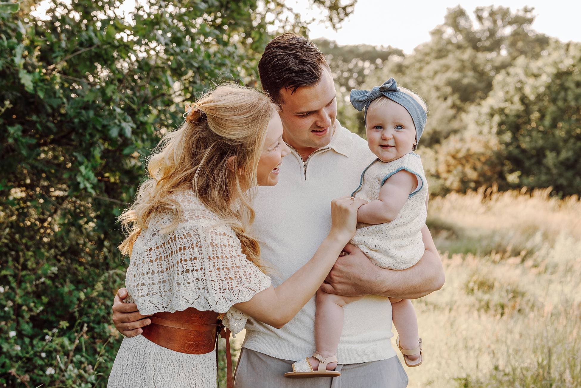 Parents cuddling toddler in tall grass during summer evening session
