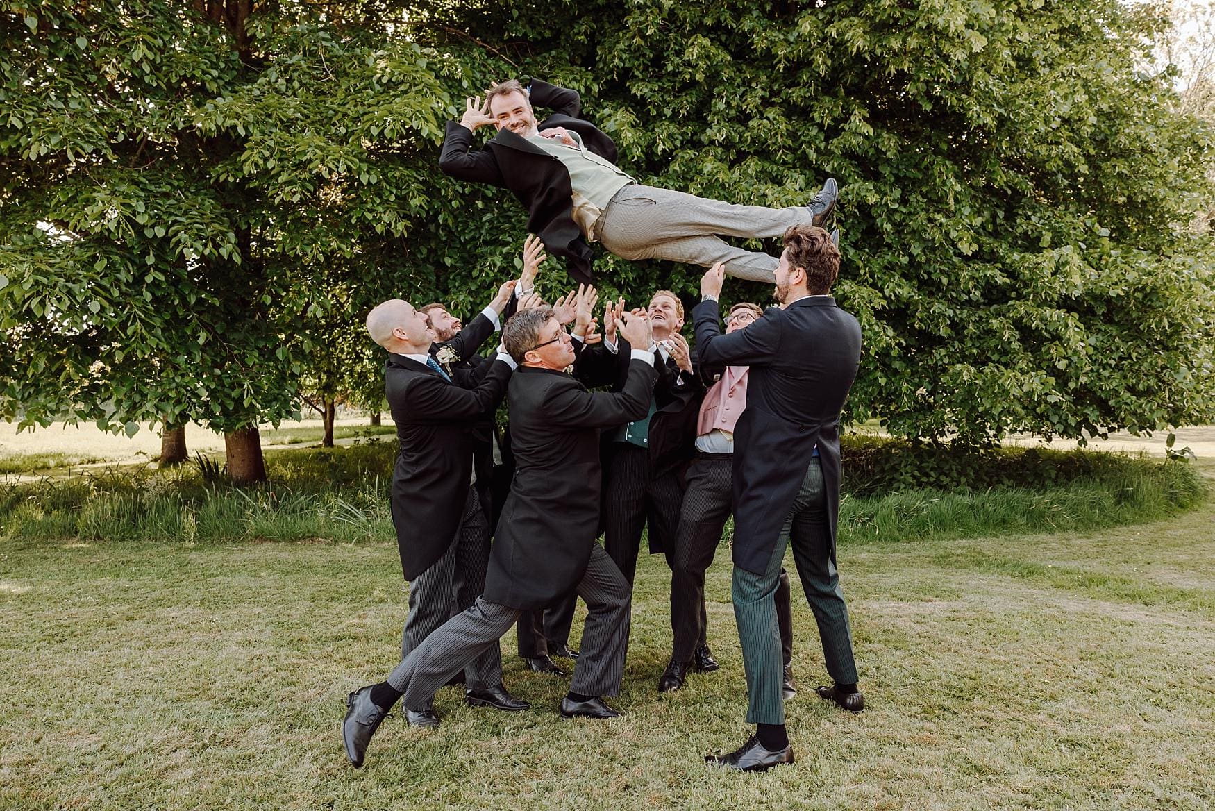 Groomsmen lifting the groom into the air on the lawn during a joyful Tithe Barn Petersfield wedding, photographed in a natural documentary style.