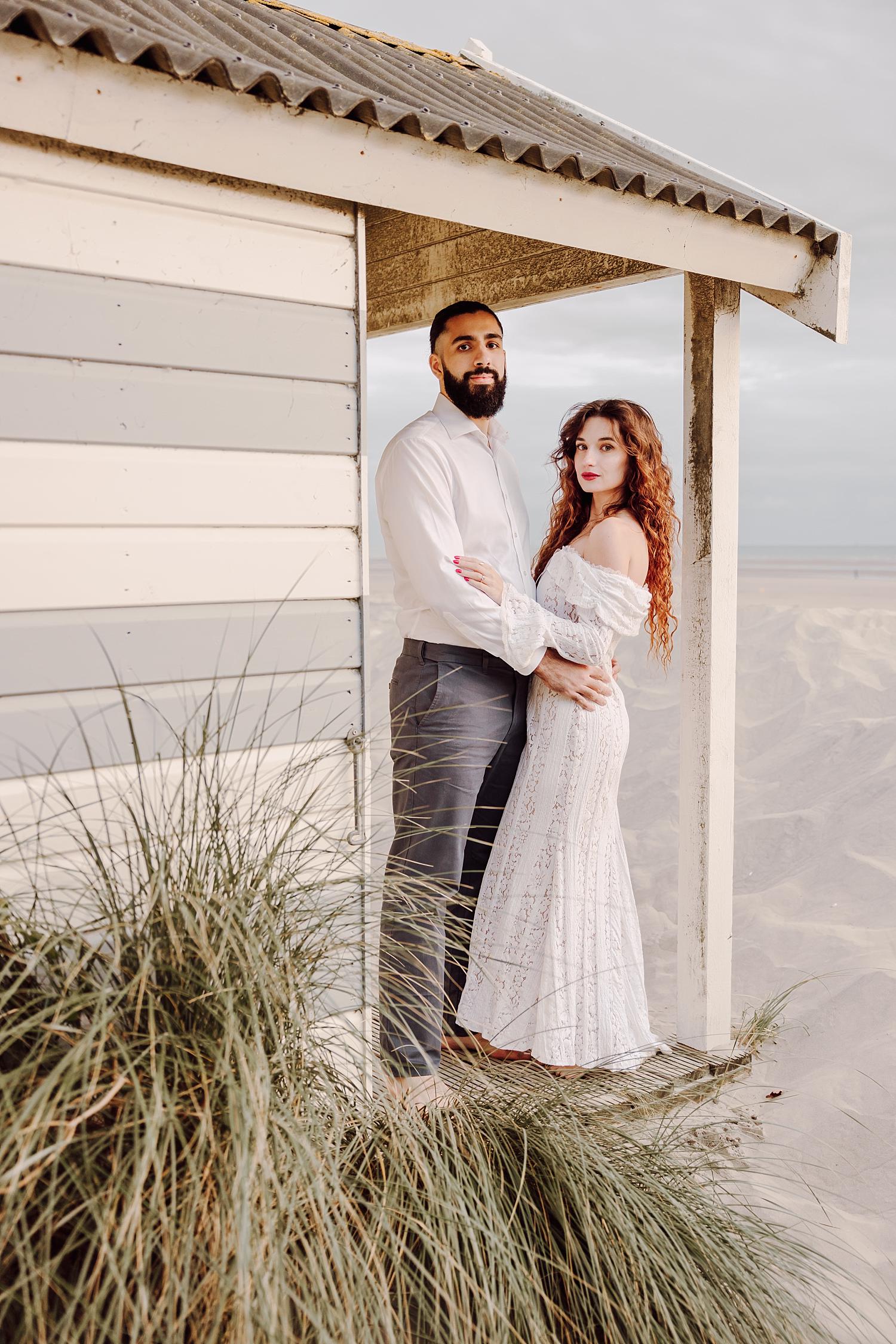 Half length engagement portrait of bride in white lace dress standing by beach hut on West Wittering dunes