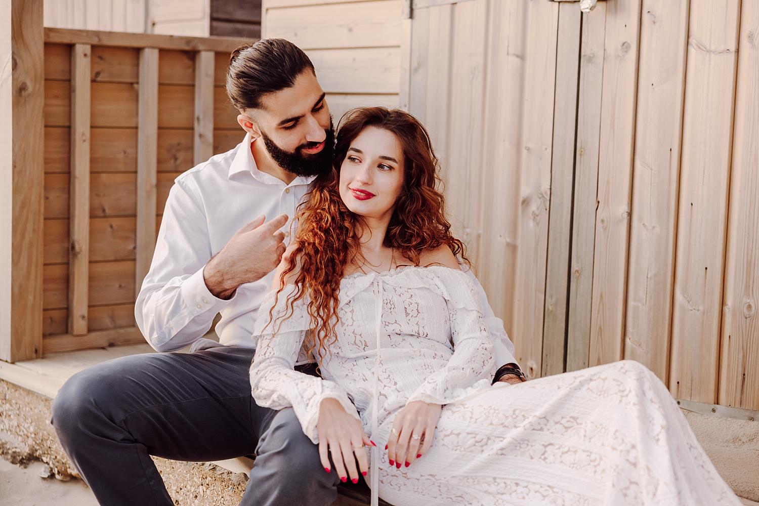 Intimate West Sussex engagement portrait of couple sitting together by wooden beach hut at sunset