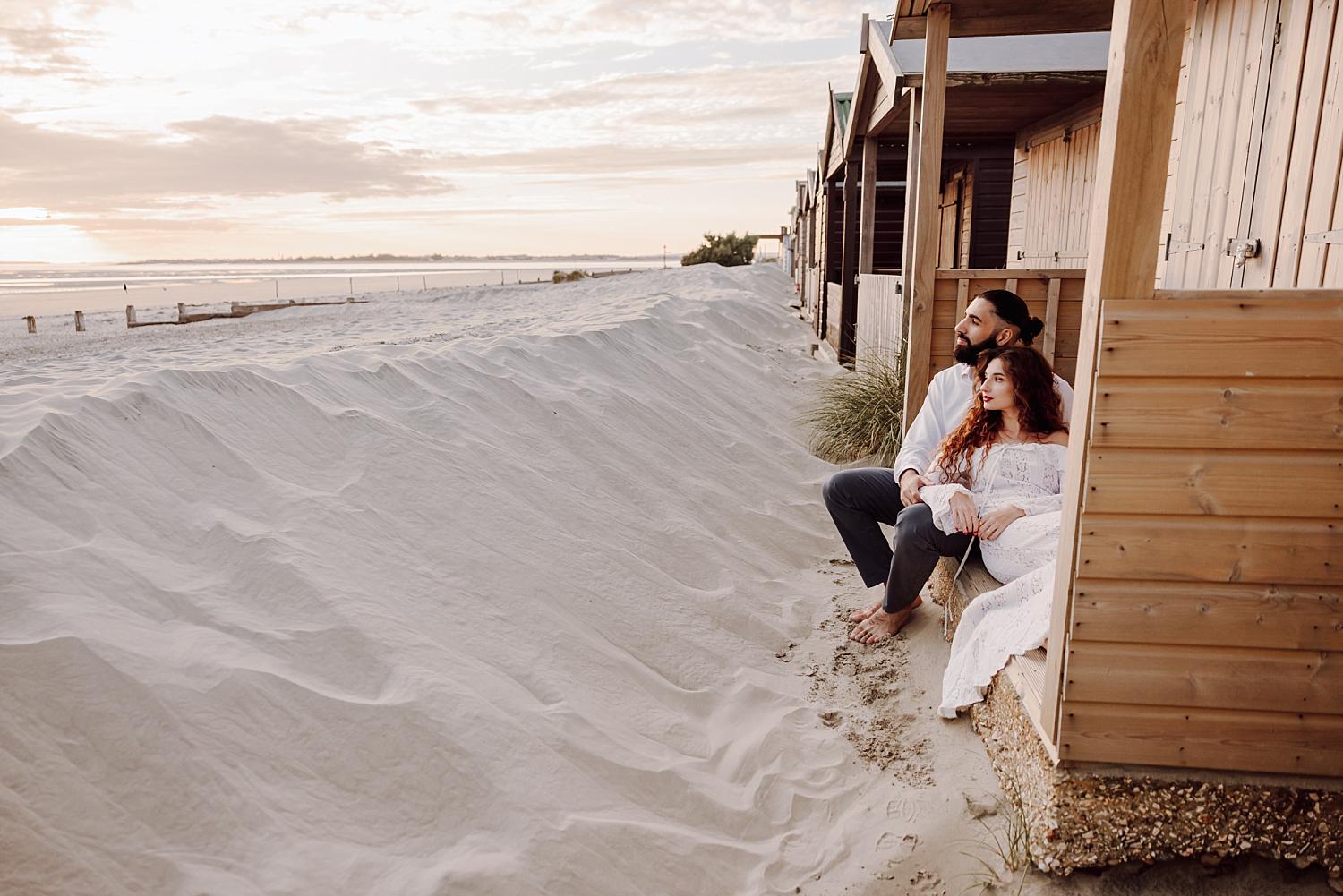 Bride sitting barefoot in sand beside beach hut during West Wittering engagement session at sunset.