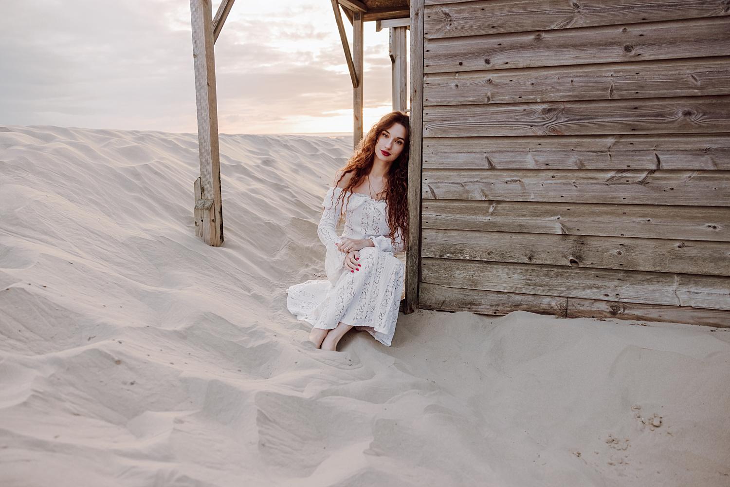 Bride sitting barefoot in sand beside wooden beach hut during sunset engagement session at West Wittering.
