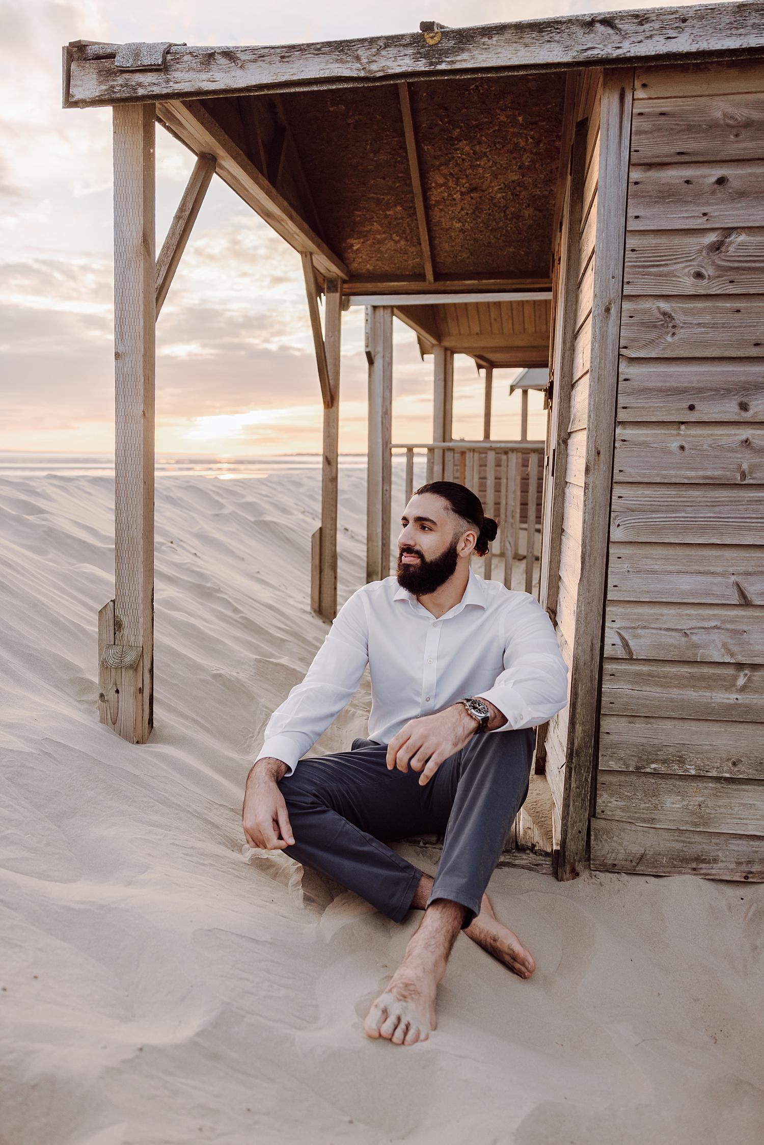 Relaxed groom sitting barefoot in sand beside beach huts at West Wittering during sunset engagement session.