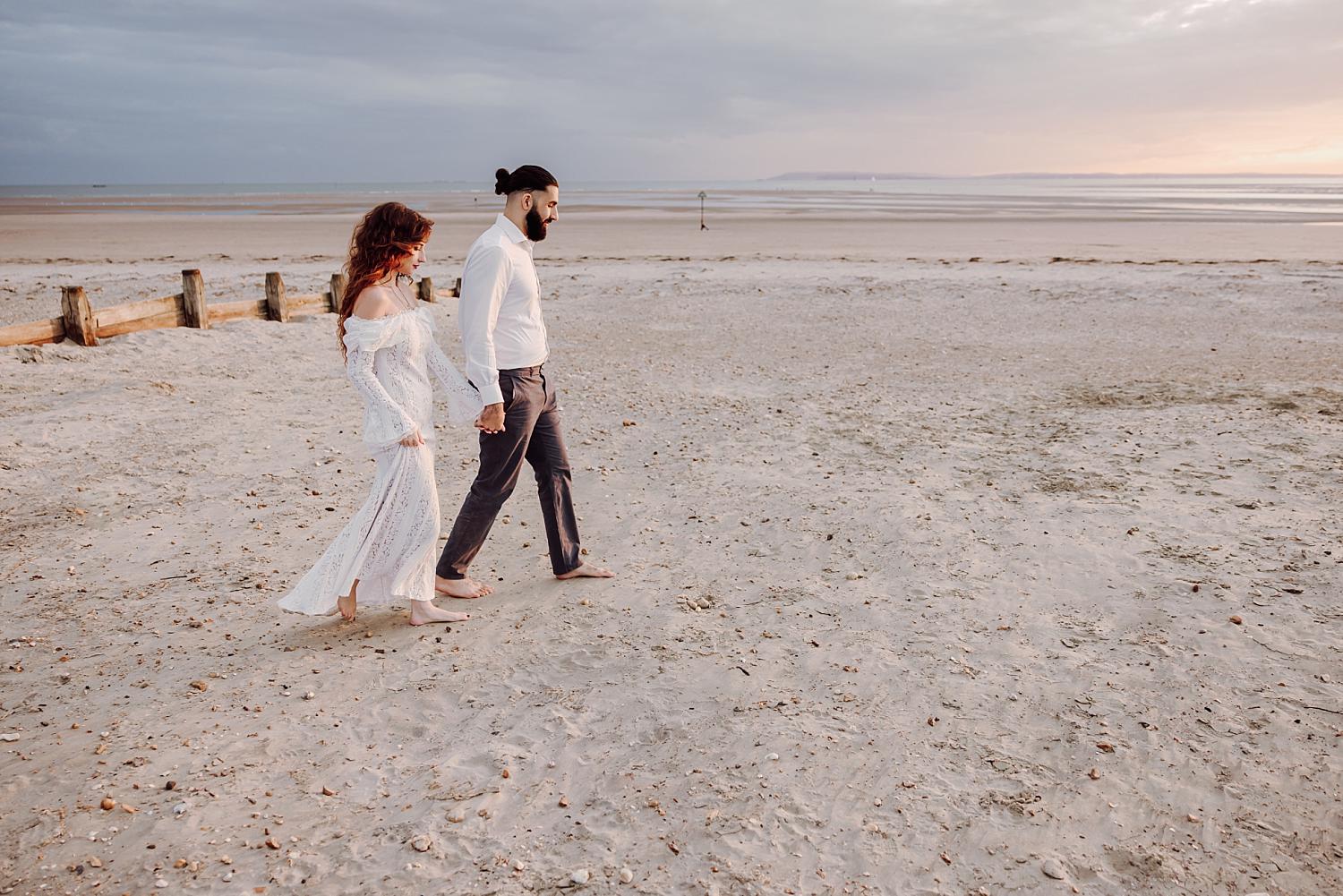 Engaged couple strolling barefoot across the sand at sunset during West Wittering beach engagement photos.