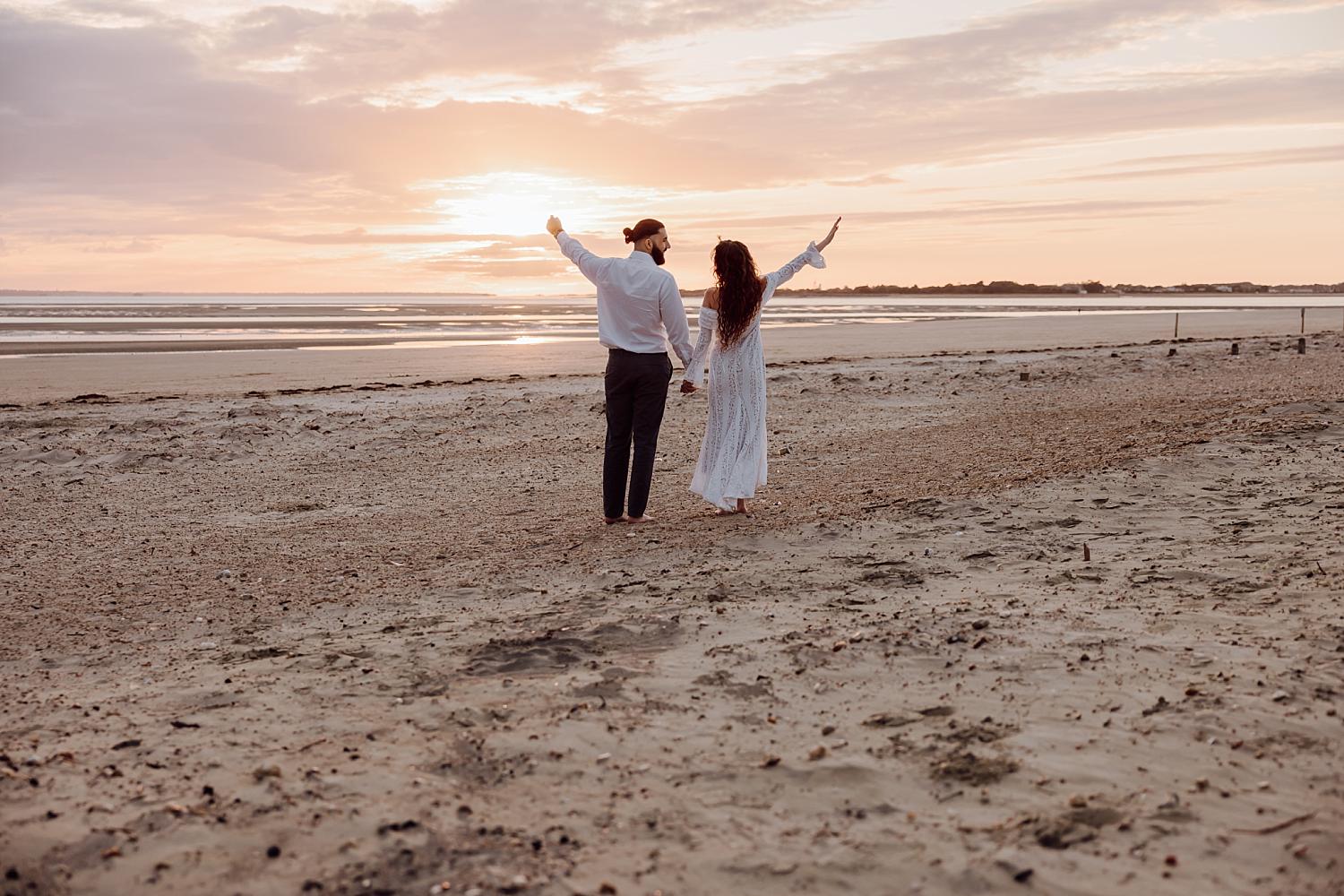 West Wittering golden hour engagement photos of couple celebrating with raised arms against warm sunset sky.