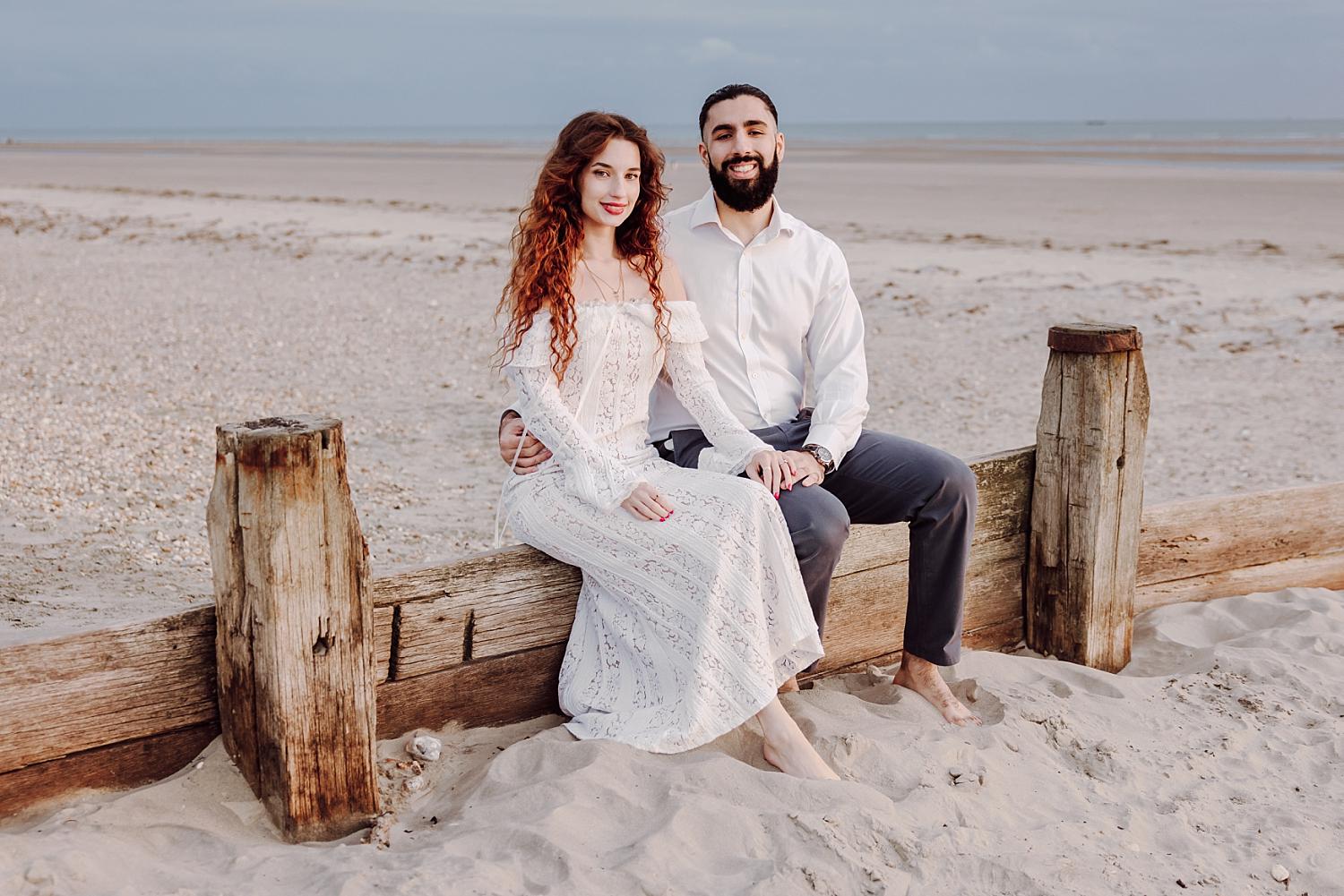 Engaged couple seated on wooden groynes at West Wittering Beach during relaxed West Sussex engagement session.