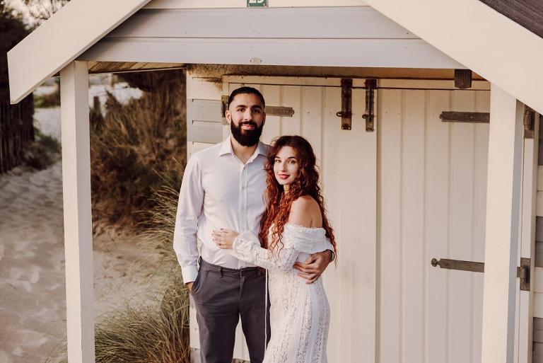 Engaged couple standing together beside beach hut at West Wittering Beach during golden hour.