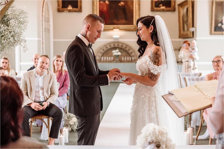 Groom placing the ring on the bride's finger beneath the mayoral portraits in the Atherley Chamber