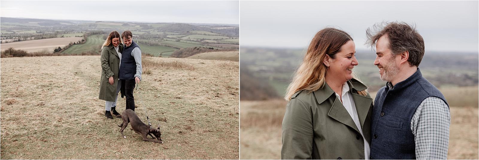Engaged couple standing with their whippet against the wide open landscape of Butser Hill