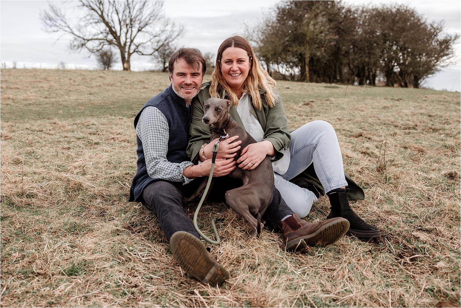 Engaged couple sitting on the grass cuddling their whippet at Butser Hill