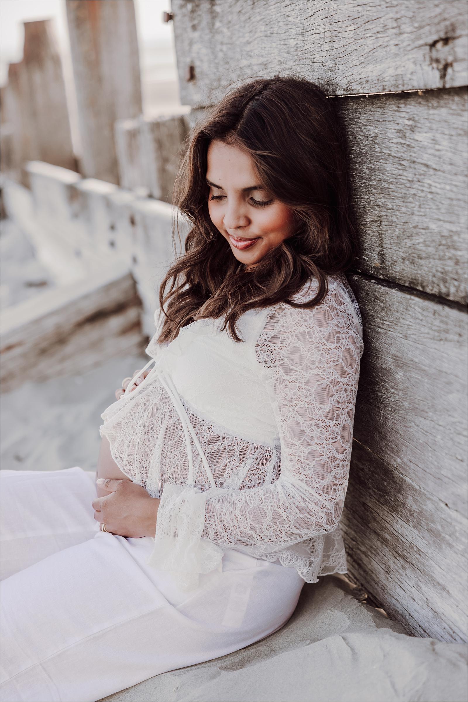 Mum-to-be in white lace resting against weathered wood in soft evening light