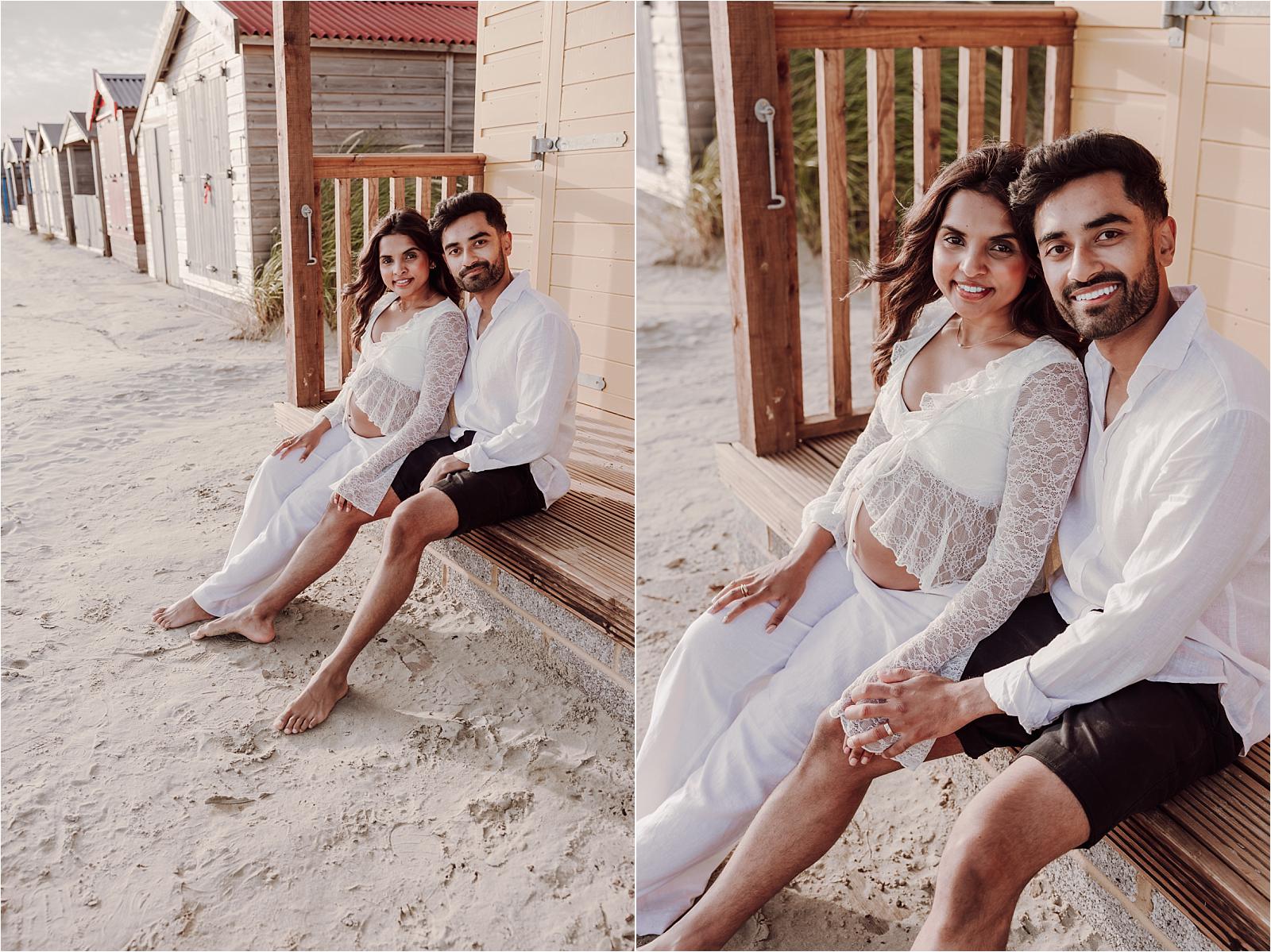 Two portraits of an expectant couple sitting on the sandy path between beach huts at West Wittering