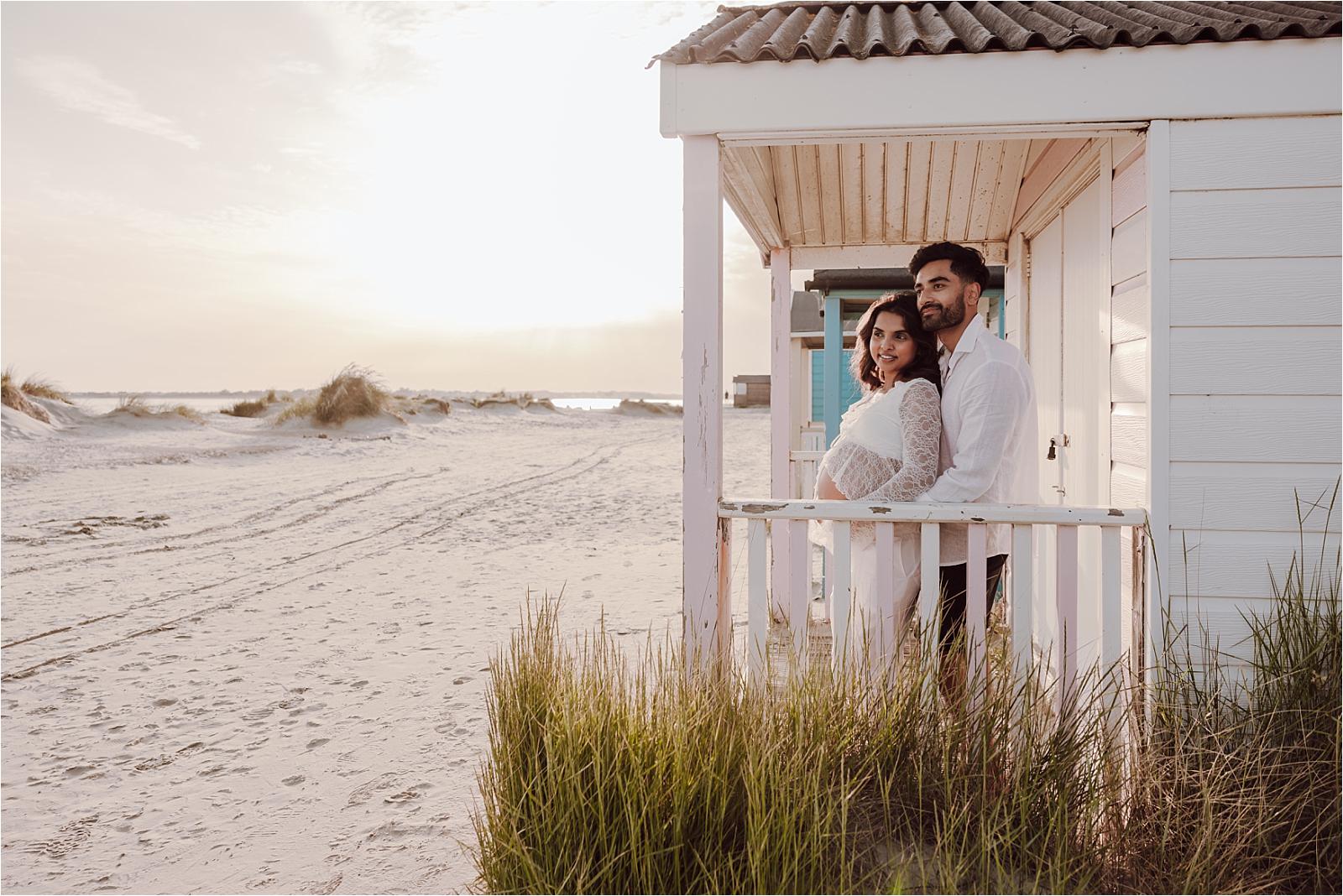 Expectant couple standing beneath a beach hut porch at sunset at West Wittering