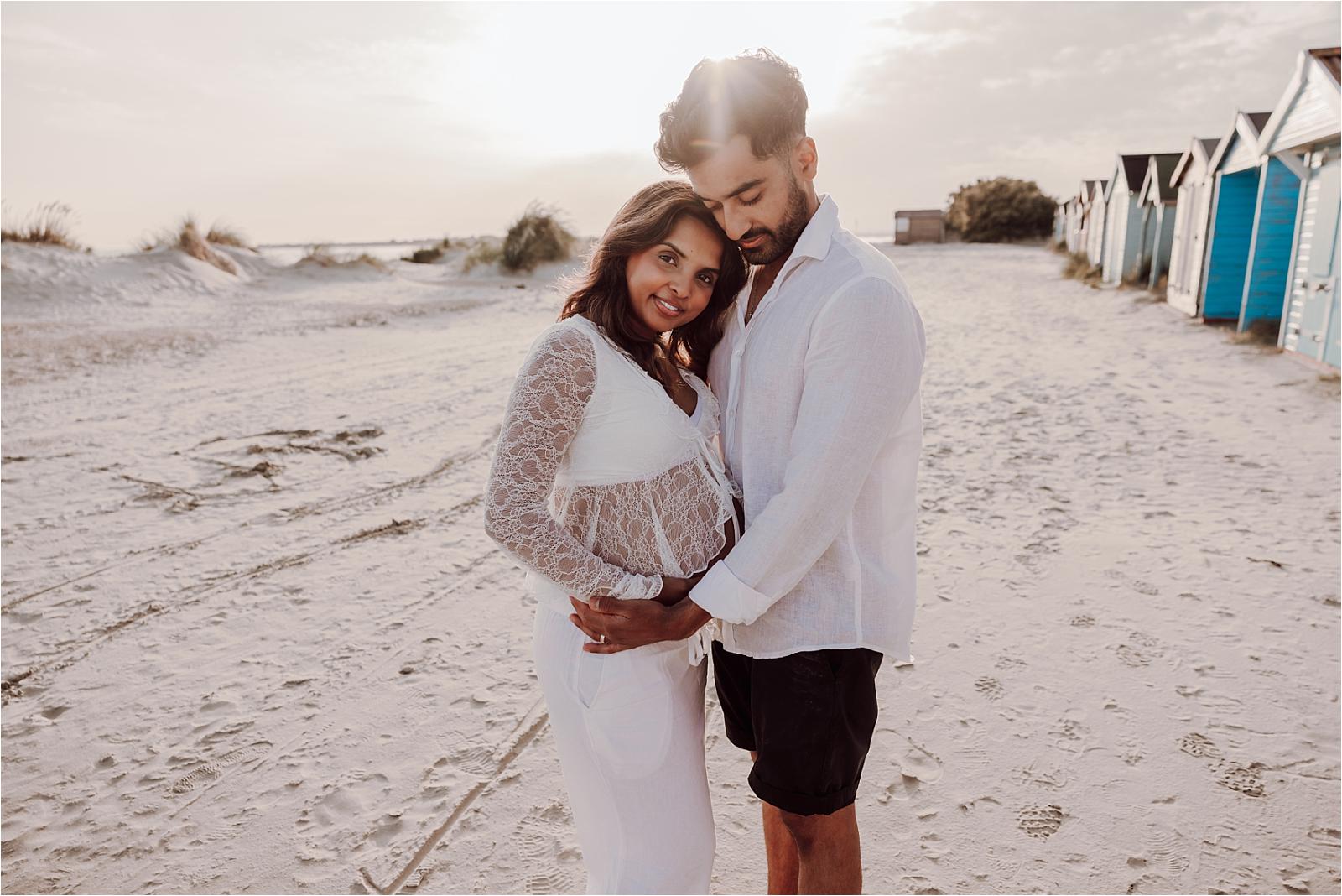 Expectant couple embracing on the sand with beach huts in the distance during their West Wittering maternity session