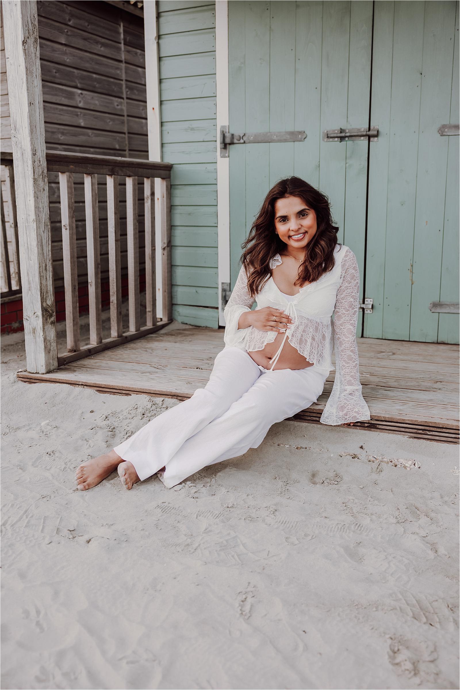 Smiling mum-to-be in white lace sitting in front of a pale blue beach hut during her maternity photoshoot at West Wittering