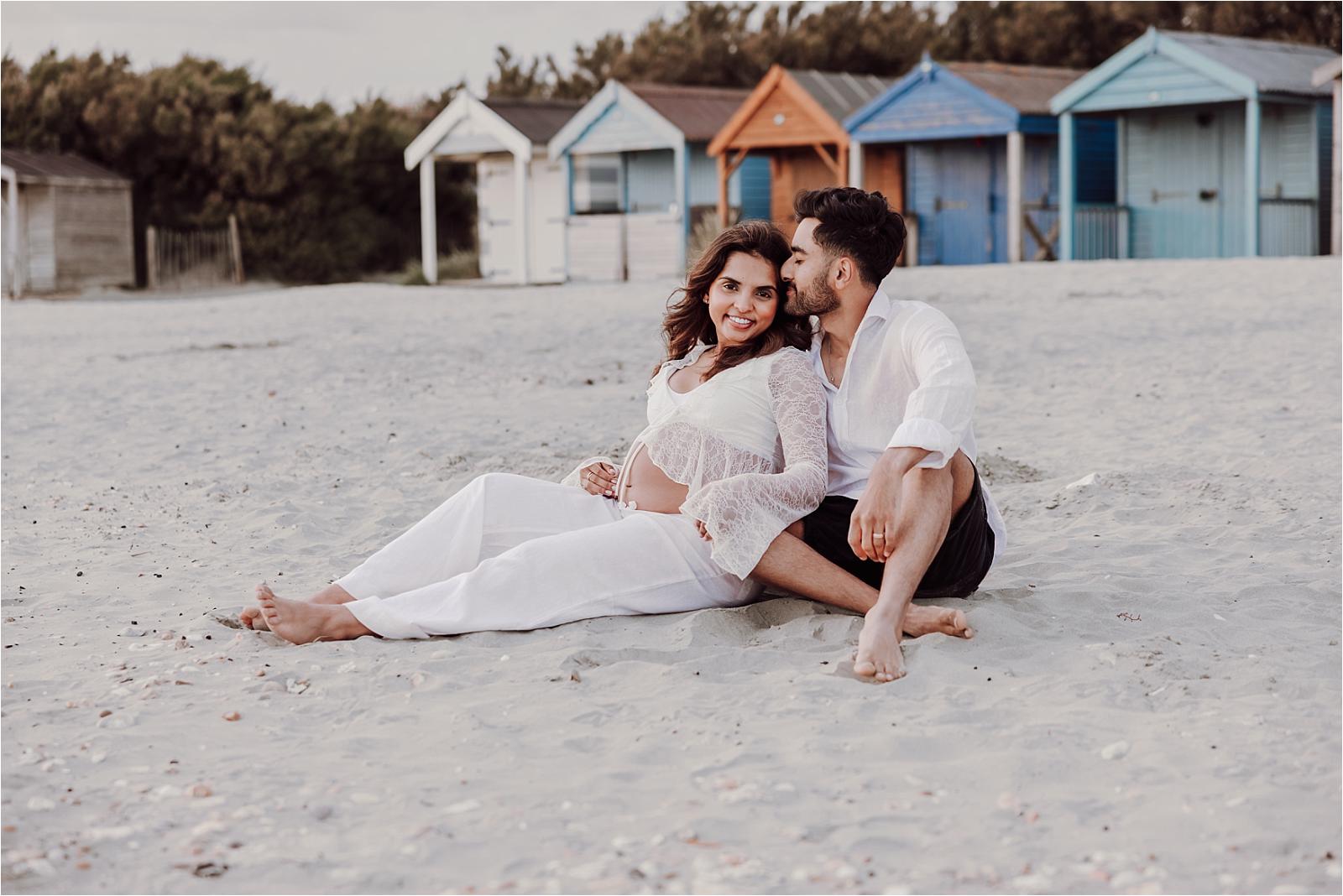 Expectant couple sitting together on the sand with beach huts behind them at West Wittering
