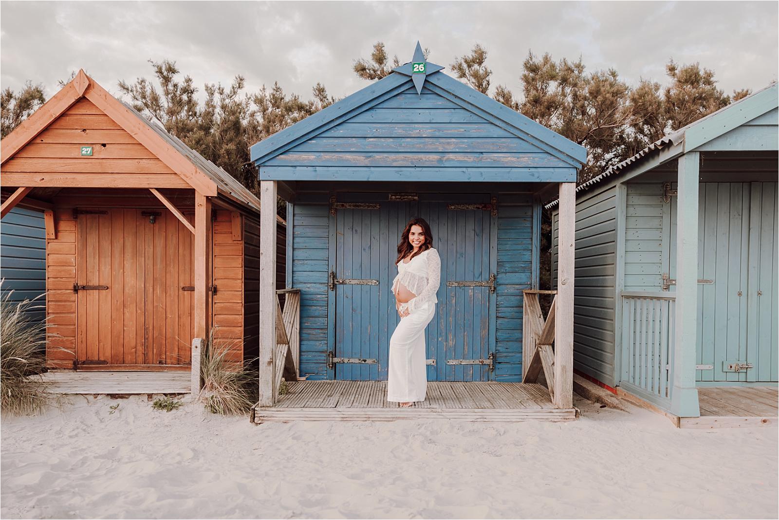 Mum-to-be in white lace beside a beach hut during her maternity photoshoot at West Wittering