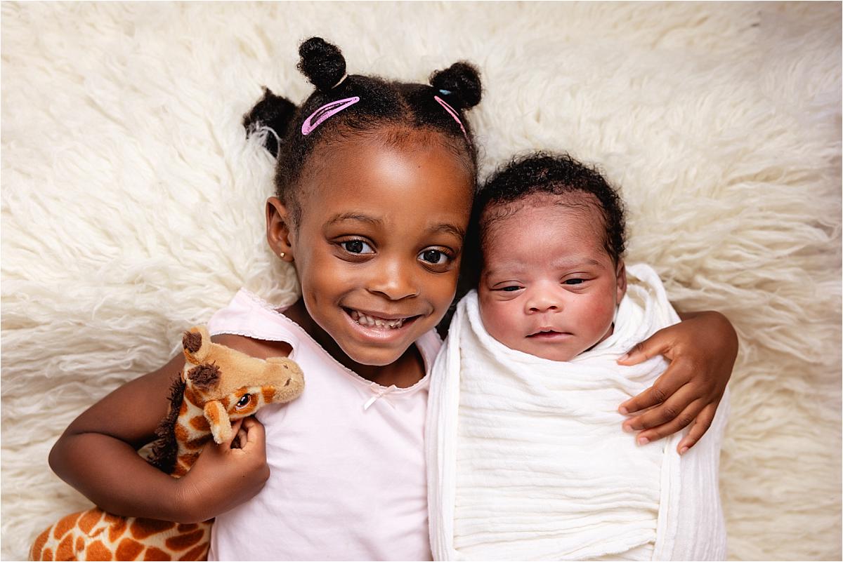 havant-newborn-with-big-sister-at-home Older sister lying beside her swaddled newborn brother holding her giraffe toy at home in Havant